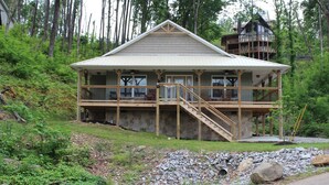 A two-story wooden house with a balcony and stairs, surrounded by trees and a rocky landscape. - Built in 2018 this is a charming, spacious chalet that sleeps 6!   (Gatlinburg)