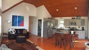 Un vaste espace de vie comprenant un canapé en cuir noir, une table basse en bois et un grand tableau accroché au mur. On aperçoit à l’arrière-plan une cuisine équipée d’appareils électroménagers modernes et un coin repas avec des chaises en bois.