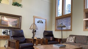 A cozy living room with a wooden coffee table, two leather armchairs, a patterned sofa, a framed picture, a lamp, and a large window with blinds.