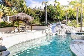 A swimming pool with a natural rock waterfall feature, surrounded by tropical vegetation and a thatched-roof structure.