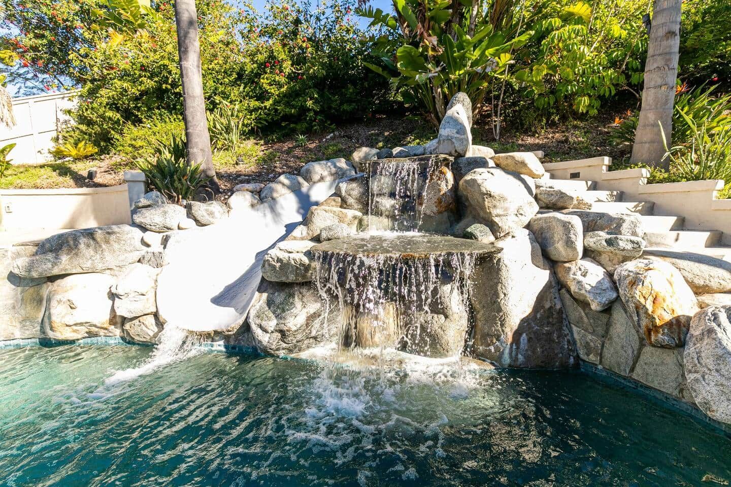 A rock waterfall feature in a landscaped pool area with surrounding vegetation.