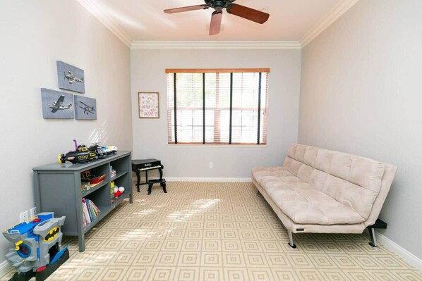 Una habitación con alfombra color arena estampada, un sofá color beige, un ventilador de techo, una ventana con persianas y una estantería con juguetes.