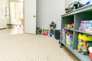 Una habitación con alfombra estampada, una estantería con juguetes y libros, y una puerta que da a otra habitación.