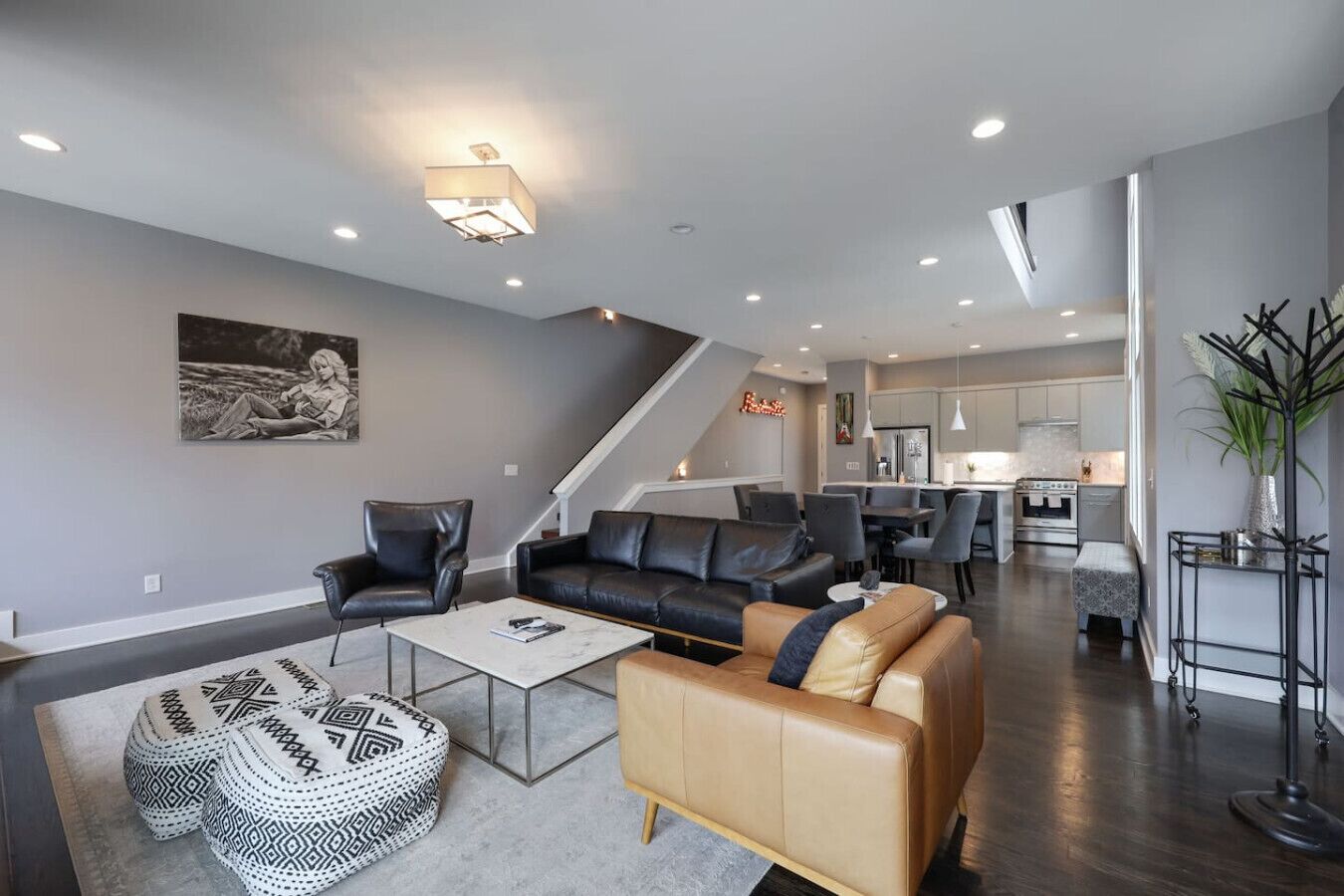 A modern living room with a sofa, armchairs, and a coffee table. A staircase leads to a kitchen and dining area.