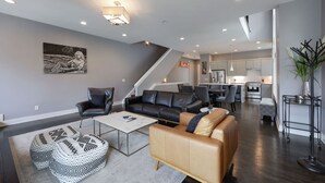 Una sala moderna con un sofá, sillones y una mesa de centro. Una escalera conduce a una cocina y un comedor.