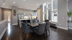 Una cocina moderna con una isla central, comedor con mesa de madera oscura y sillas tapizadas en gris, y amplios ventanales con persianas.