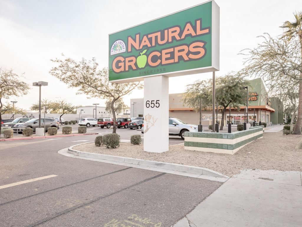 Een bord van Natural Grocers met een kleurencombinatie van groen en oranje, gelegen op de hoek van een straat met een parkeerplaats en een gebouw op de achtergrond.