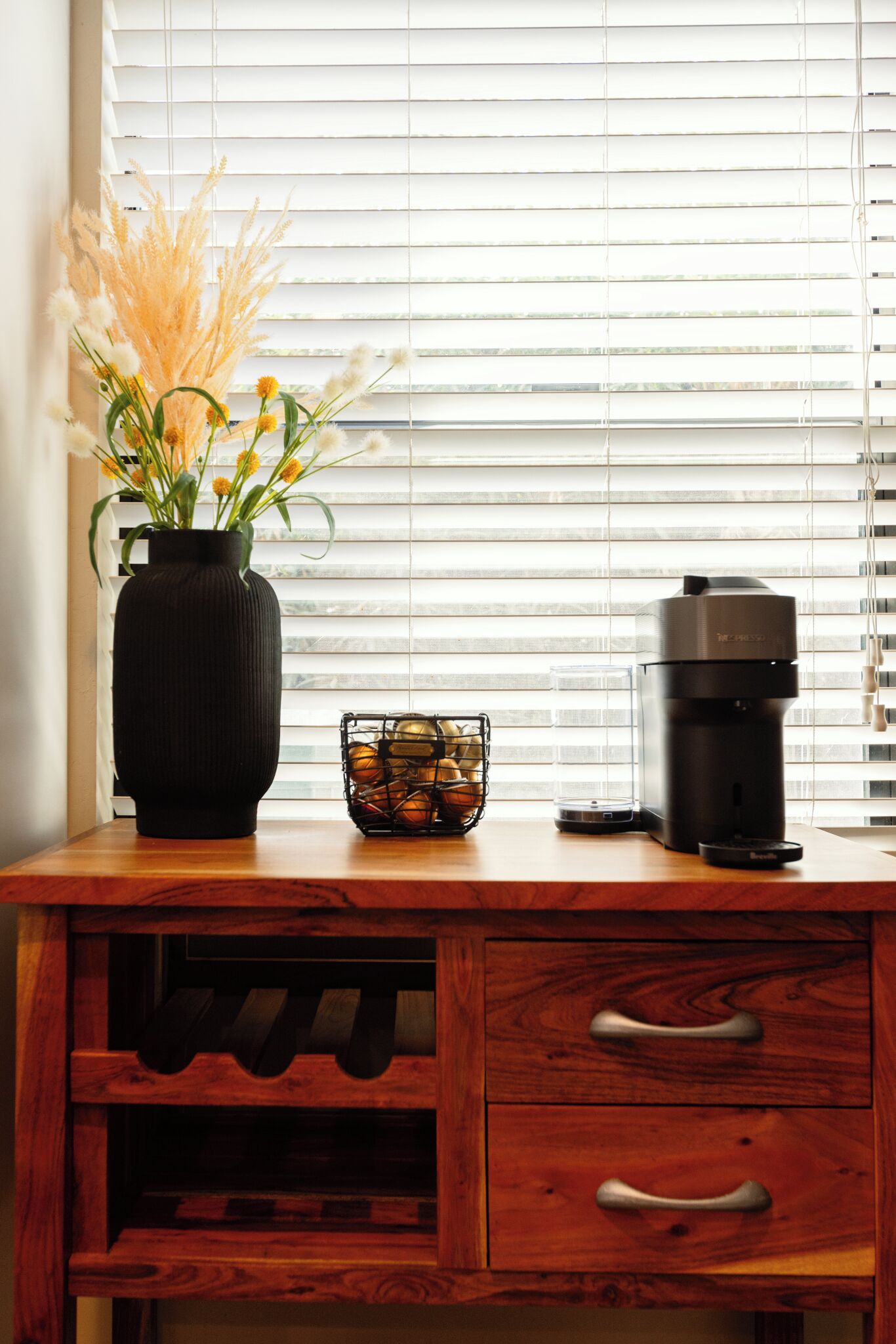 Un escritorio de madera con un florero negro de flores, una cesta de naranjas y una cafetera.