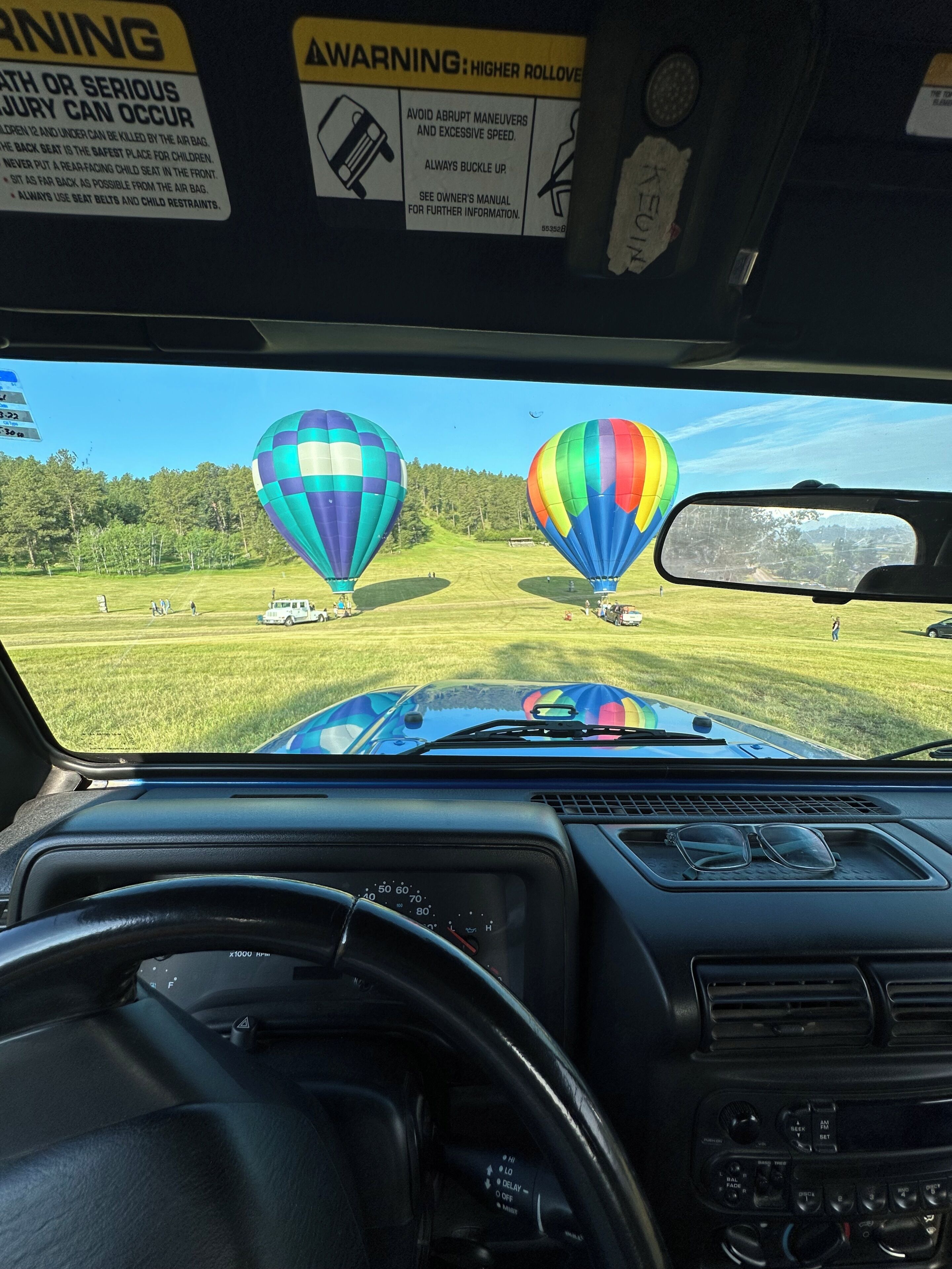 A car's interior with a view of two colorful hot air balloons on a grassy field.