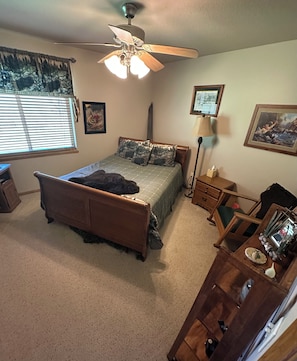 A bedroom with a bed, a ceiling fan, a window with blinds, a bedside table, a lamp, and a framed picture on the wall.
