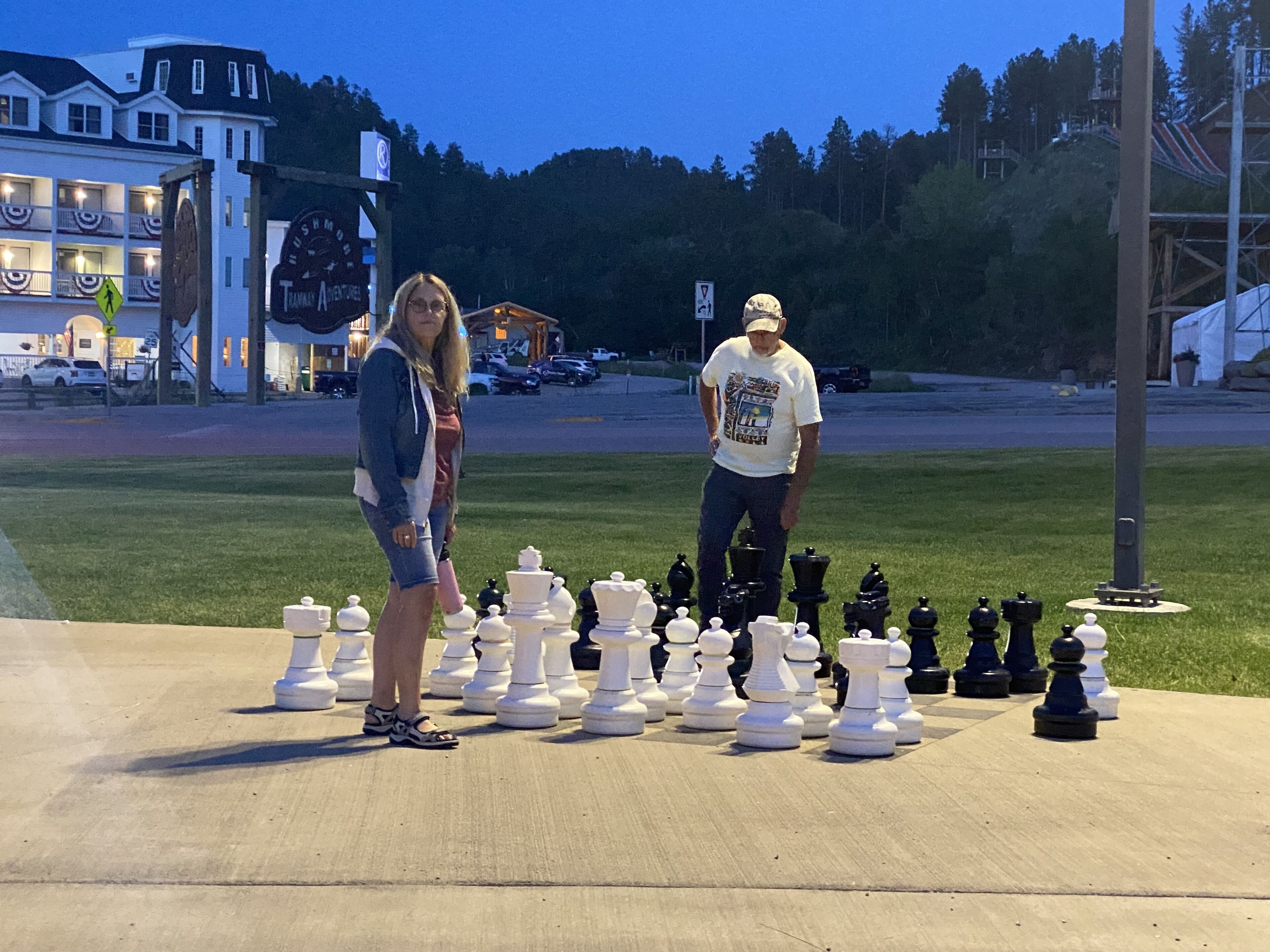Un jeu d’échecs est installé sur une surface en béton à l’extérieur, et deux personnes se tiennent à côté.