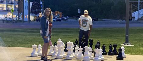 Un jeu d’échecs est installé sur une surface en béton à l’extérieur, et deux personnes se tiennent à côté.