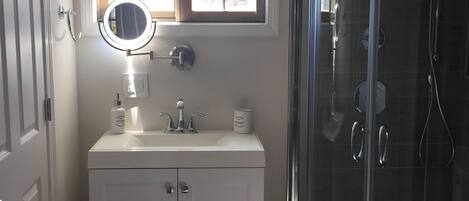 Un bagno con un mobile vanitario bianco, uno specchio rotondo, una finestra sopra il lavandino e una cabina doccia in vetro.