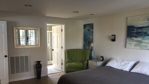 Un dormitorio con cama, una silla, una mesita de noche, un cuadro y una puerta que da a otra habitación.