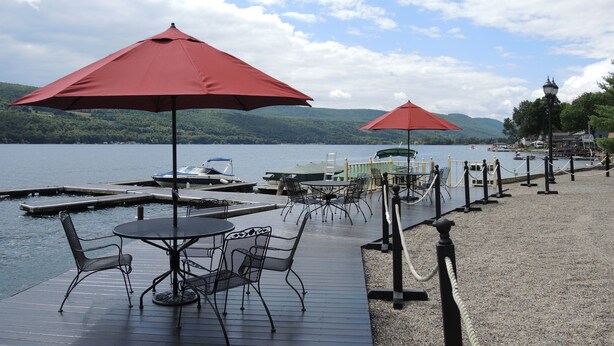 Terraza junto al lago con mobiliario exterior y sombrillas, embarcaciones en el muelle, y vistas al lago y las colinas.