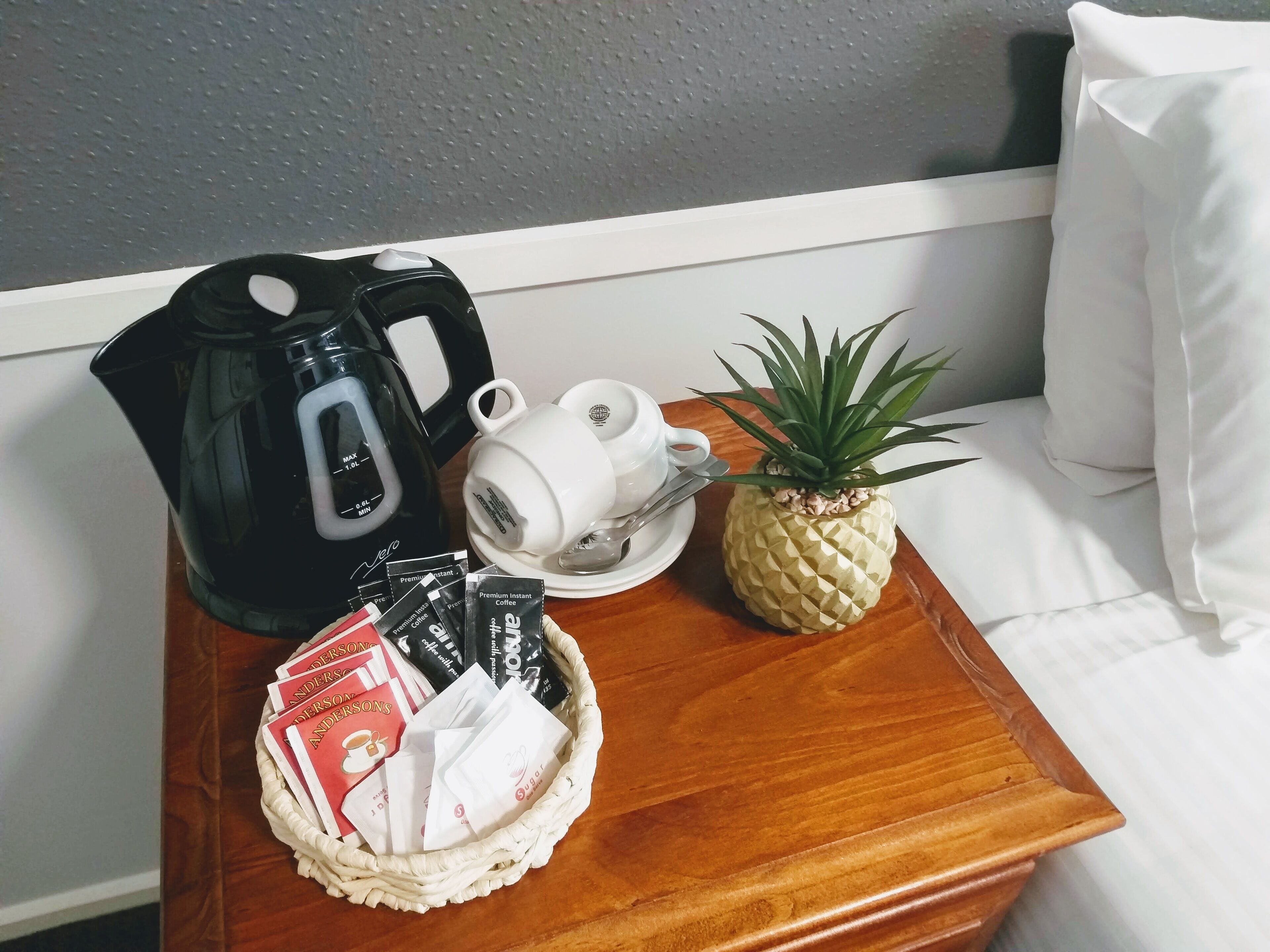 Een houten bijzettafeltje met een zwarte koffiezetapparaat, een witte mok, een ananas en een mandje met koffieporties.