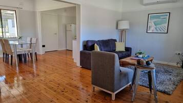 A spacious living area with a wooden floor, a grey armchair, a sofa with cushions, a small table with decorative items, and a painting on the wall.