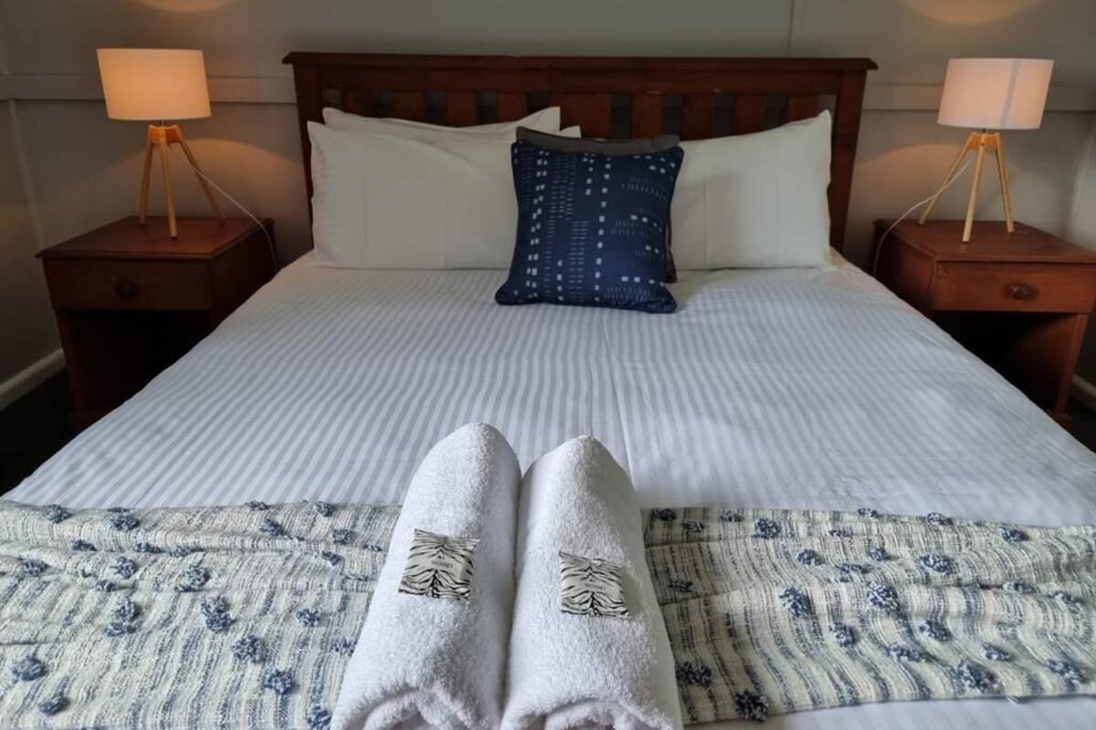Ein ordentlich bezogenes Bett mit weißen Leinen, einem blauen Kissen mit Muster und zwei Nachttischlampen auf Holznachttischen.