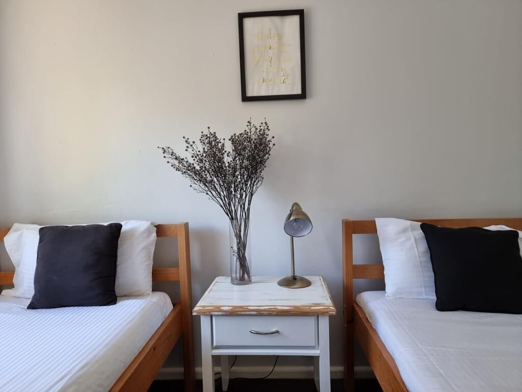 Une chambre avec deux lits simples, chacun doté d’une tête de lit en bois et de linge de lit blanc. On y trouve une petite table de chevet avec une lampe et un vase contenant des fleurs séchées. Un tableau encadré est accroché au mur, au-dessus des lits.