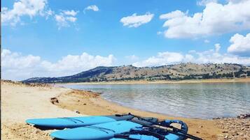 Una playa con un kayak y una pala sobre la arena, un río tranquilo al fondo y colinas en la distancia.