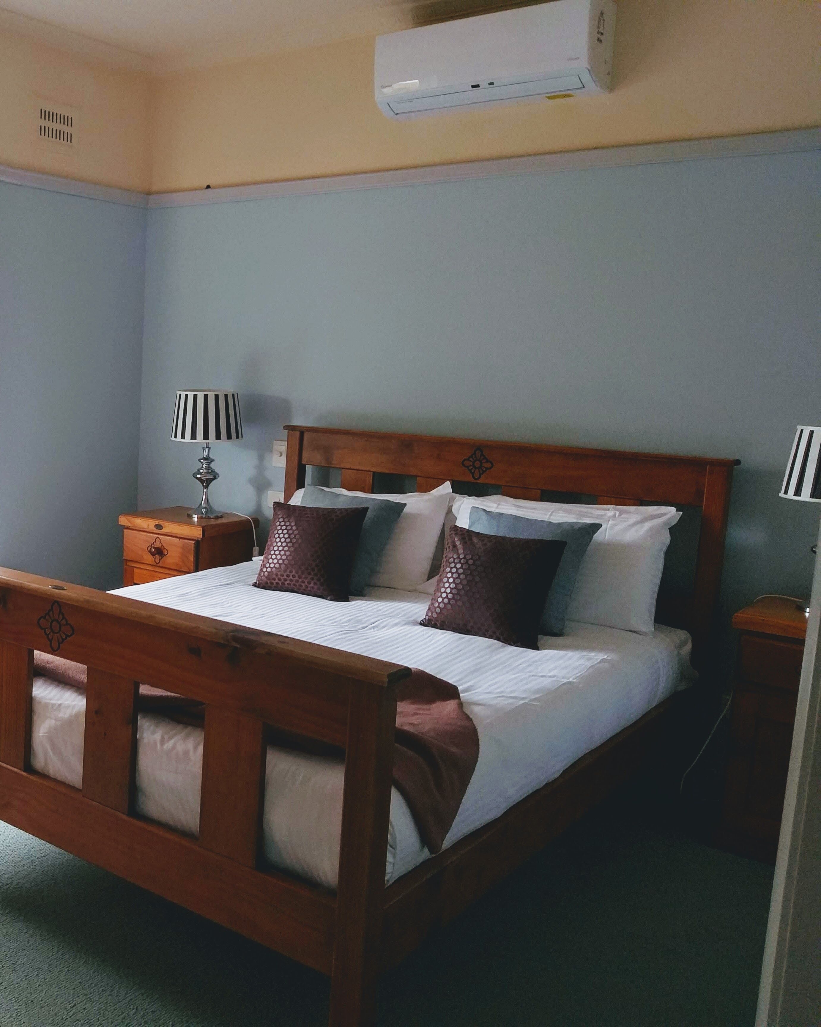 A wooden bed with a headboard, a bedside table, a lamp, and a wall-mounted air conditioner.