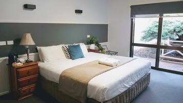 Cama bem arrumada com manta e travesseiros bege, mesa de cabeceira com abajur, uma pequena planta e janela com vista para árvores.