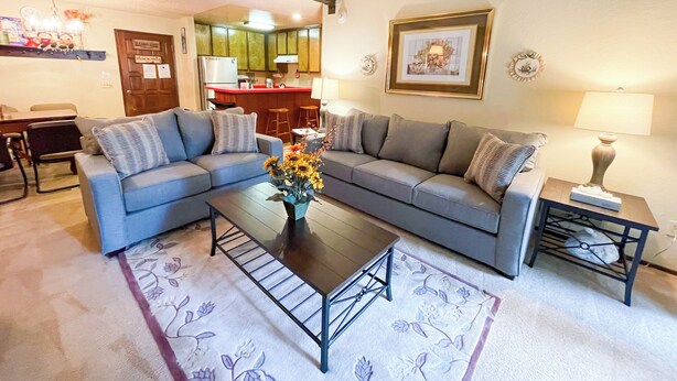 Ein Wohnzimmer mit grauem Sofa, einem Couchtisch und einem Teppich mit Blumenmuster. Im Hintergrund ist eine Küche mit einer roten Arbeitsplatte und hölzernen Barhocker zu sehen.
