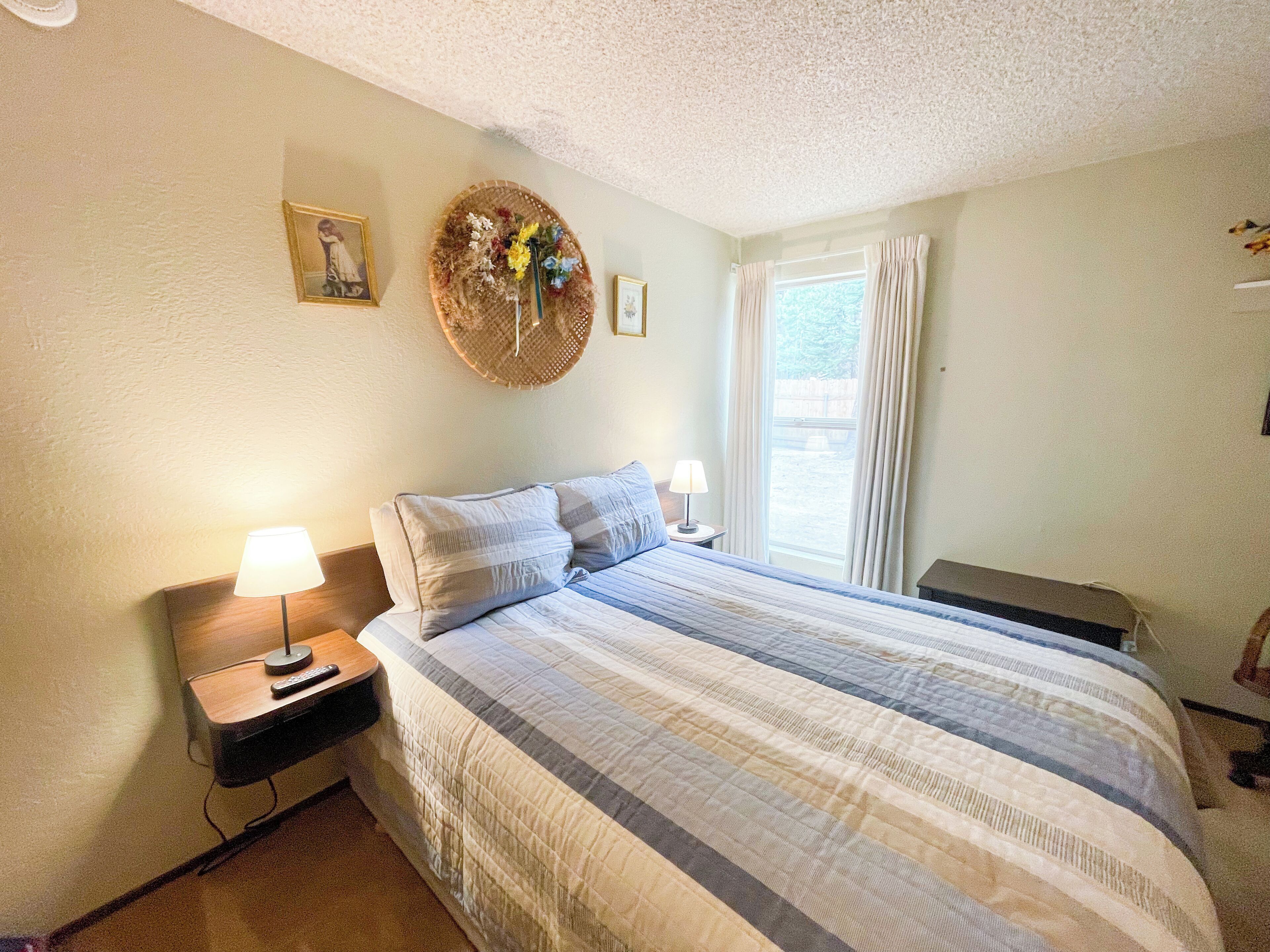 Un dormitorio con una cama grande, dos mesitas de noche, una lámpara, una cesta en la pared con flores y una ventana con cortinas.