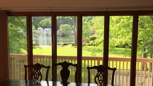 A dining area with a large table and chairs, a view of a green park with a lake and houses, and a wooden deck.