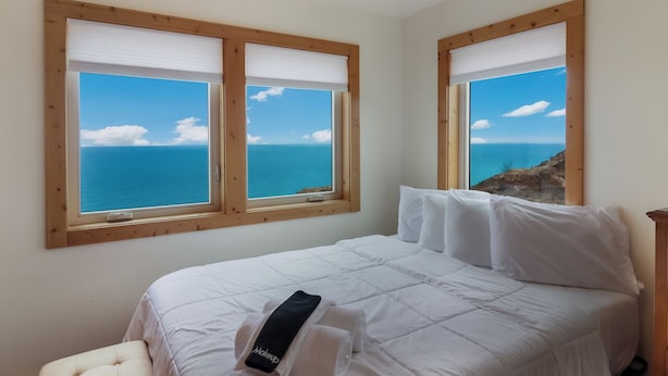 Un dormitorio con cama, dos ventanas vistas al mar y un taburete pequeño.