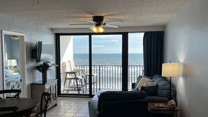 Una stanza con balcone vista mare, ventilatore a soffitto, TV, tavolo da pranzo e divano.