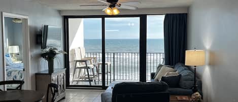 Quarto com varanda vista para o mar, ventoinha no teto, televisão, mesa de jantar e sofá.