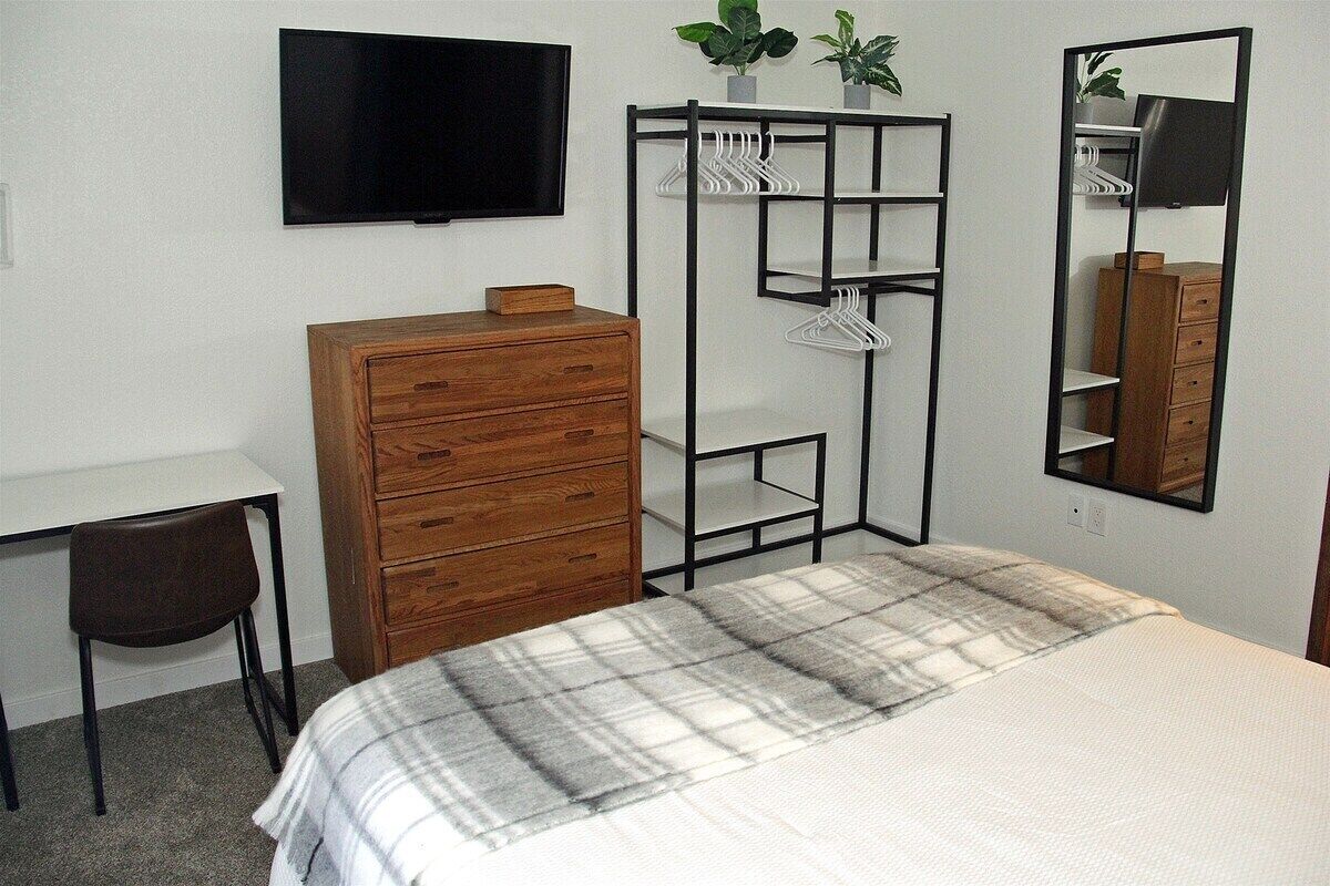 Quarto com cama, cómoda de madeira, escrivaninha, cadeira, televisão, guarda-roupa e espelho.