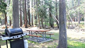 A picnic setup with a grill and table in a wooded area. - Sunshine Village 165- Centrally Located, Near Eagl (Mammoth Lakes)