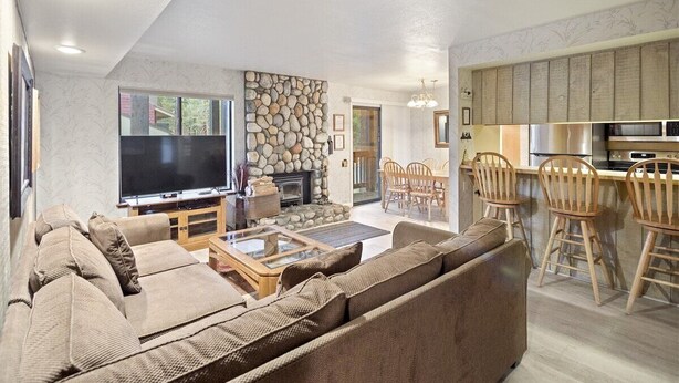 A living room with a large flat-screen TV, a stone wall, a fireplace, and a dining area with wooden chairs. - Sunshine Village 165- Centrally Located, Near Eagl (Mammoth Lakes)
