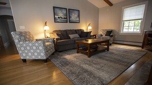 A living room with a sofa, two armchairs, a coffee table, and a rug. There are two wall-mounted lamps, a window with blinds, and two framed pictures on the wall.