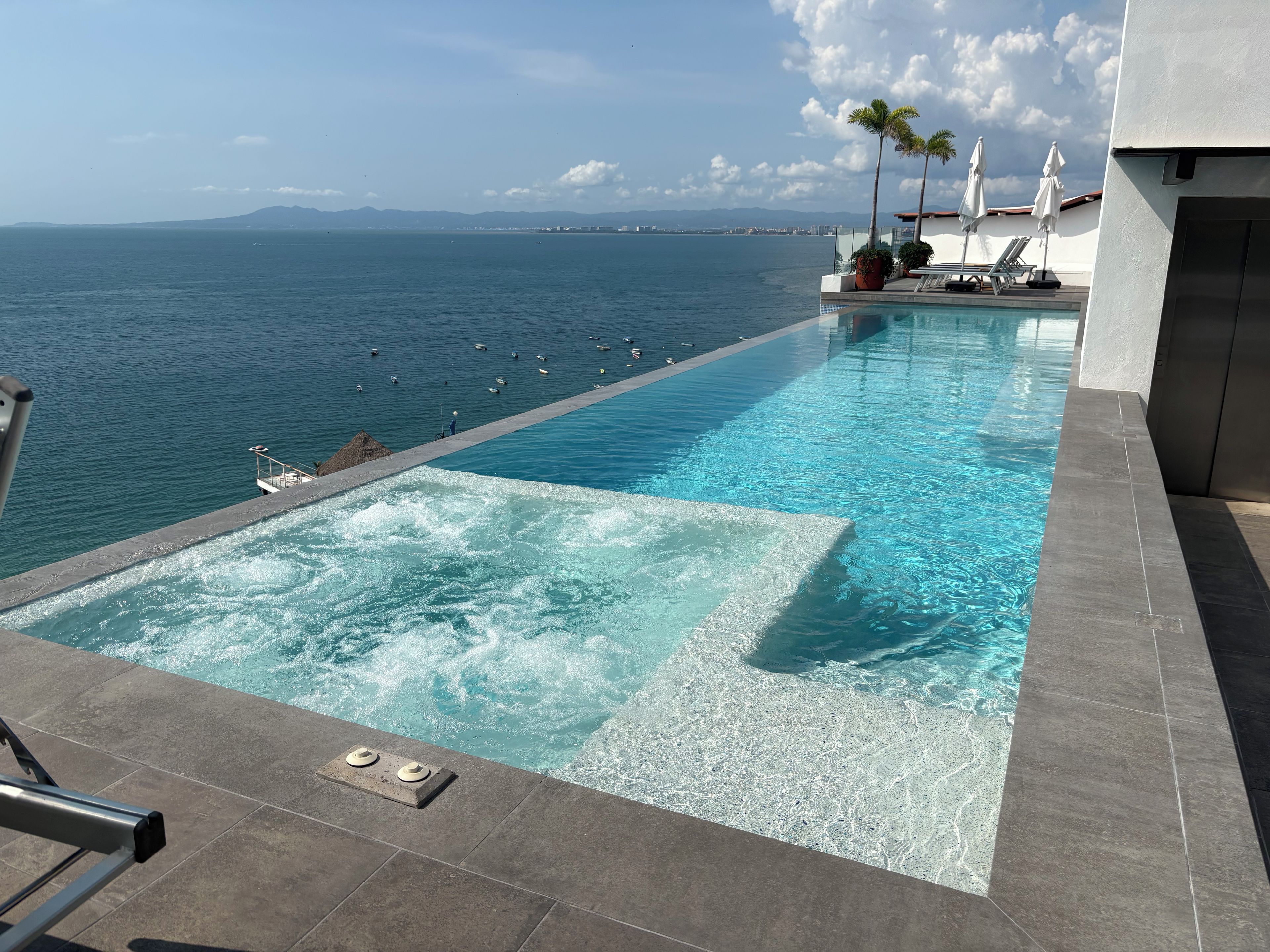 Une piscine avec vue sur la mer et les montagnes, un espace détente avec des chaises et des parasols, et un jacuzzi.