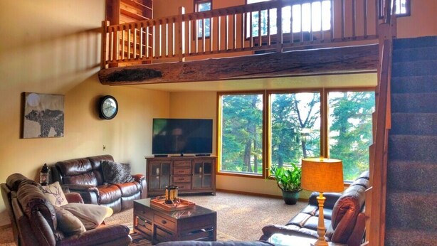 A spacious living room with a high ceiling, a wooden balcony, a large window with a view of trees, a television, and leather sofas. - Bear Paw Ranch Vacation Rentals (Bigfork)