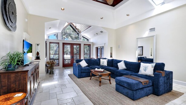 A spacious living room with a large sectional sofa, a wooden coffee table, and a TV on a wooden cabinet. There is a staircase leading to an upper level with a balcony. - SERENITY - Luxury Captiva Home, 4 Homes From the Beach (Captiva)