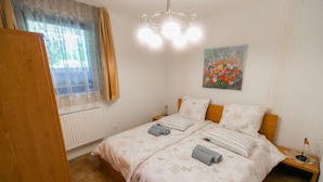Une chambre à coucher avec un lit double, deux tables de chevet, un radiateur, une fenêtre avec des rideaux et un tableau au mur.