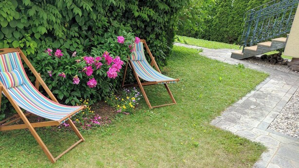 Deux fauteuils de jardin en bois, recouverts d’un tissu à rayures, sont placés sur un chemin de pierres, avec une haie verdoyante en arrière-plan.