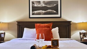 A neatly made bed with white linens, two pillows, a glass of wine, and a tray with snacks. A framed welcome sign is on the bed. - Outdoor Pool, 3 Hot tubs, Gym. Warm and Welcoming (Canmore)