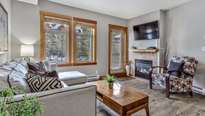 A modern living room with a sofa, a wooden coffee table, a patterned armchair, a fireplace, and a television mounted on the wall. - Outdoor Pool, 3 Hot tubs, Gym. Warm and Welcoming (Canmore)