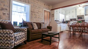 A living room with a brown sofa, patterned armchairs, and a coffee table. There is a kitchen area with white cabinets and a dining table with chairs.
