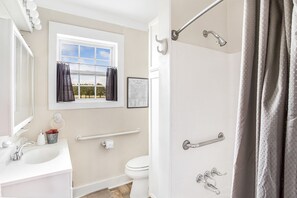A bathroom with a sink, toilet, and shower. There is a window with curtains, a towel rack, and a framed word search puzzle on the wall.