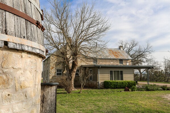 A stone building with a metal structure attached to it, a house with a metal roof, and a tree in the foreground.