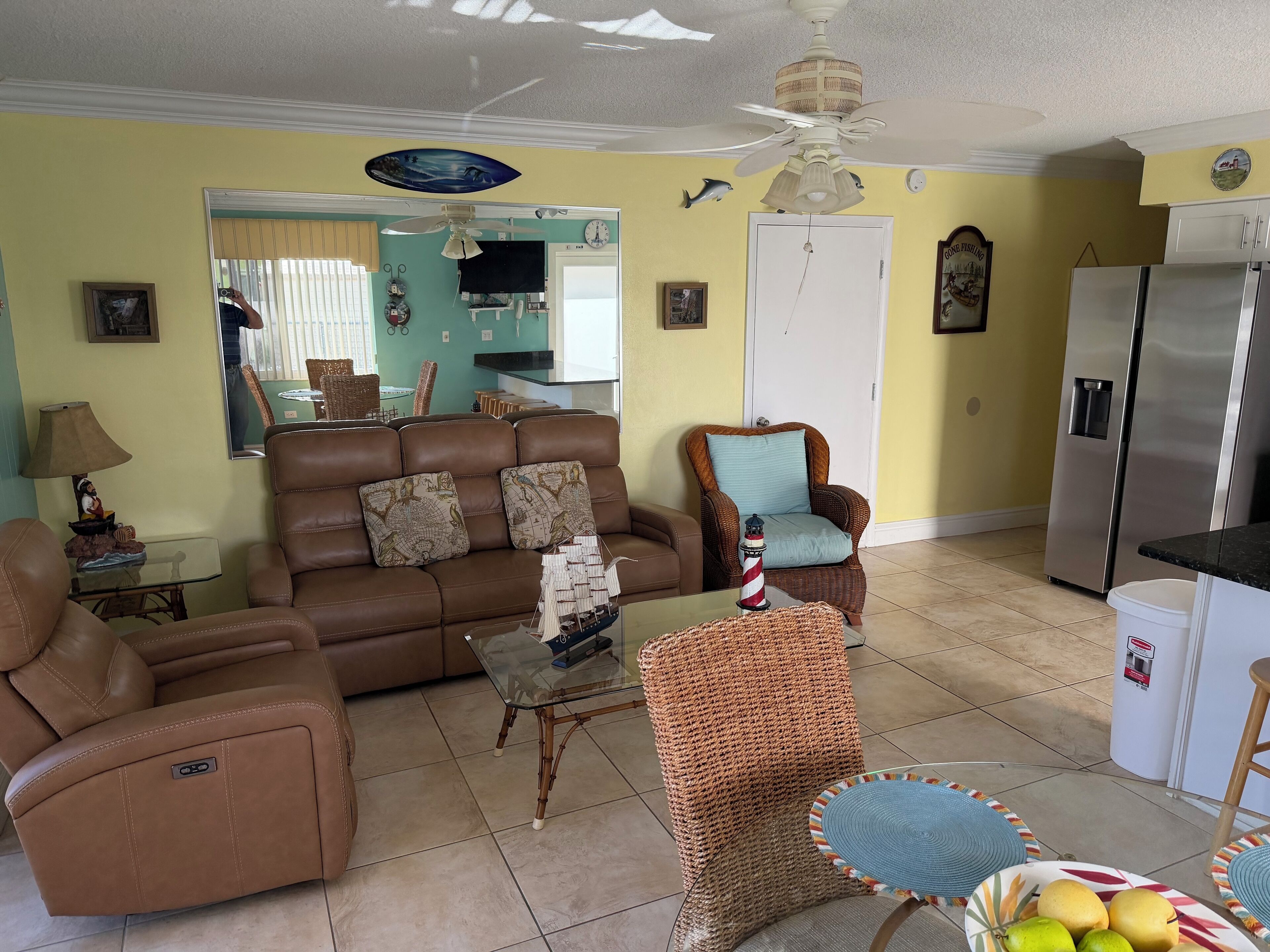 A living room with a brown leather sofa, a wicker armchair, and a glass coffee table. There is a refrigerator and a dining table in the background.