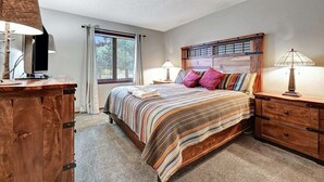 Quarto com cama grande, cabeceira de madeira, duas mesas de cabeceira com luminárias, cômoda de madeira e janela com cortinas.