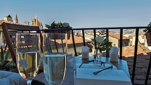 A table with two glasses of white wine, a book, and candles on a balcony overlooking a cityscape.