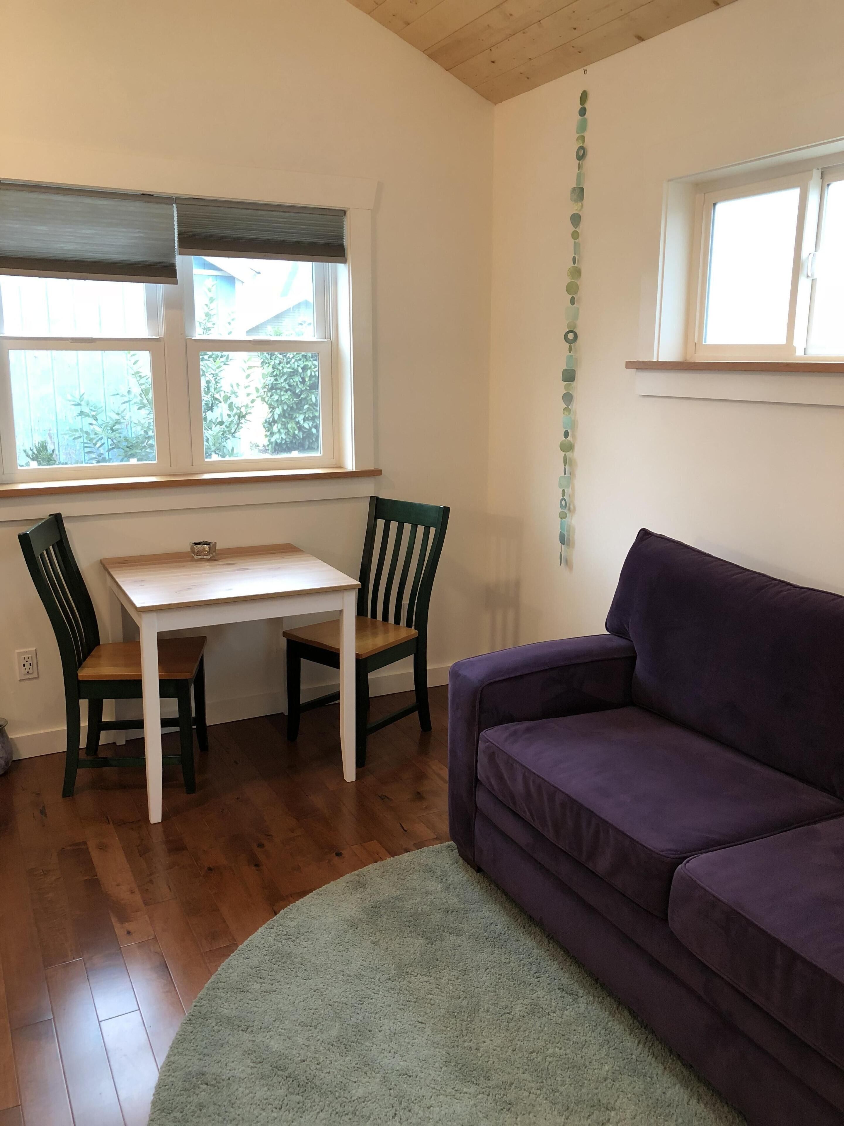 Een kamer met een houten vloer, een witte tafel met twee stoelen, een paarse bank en een raam met zonwering.